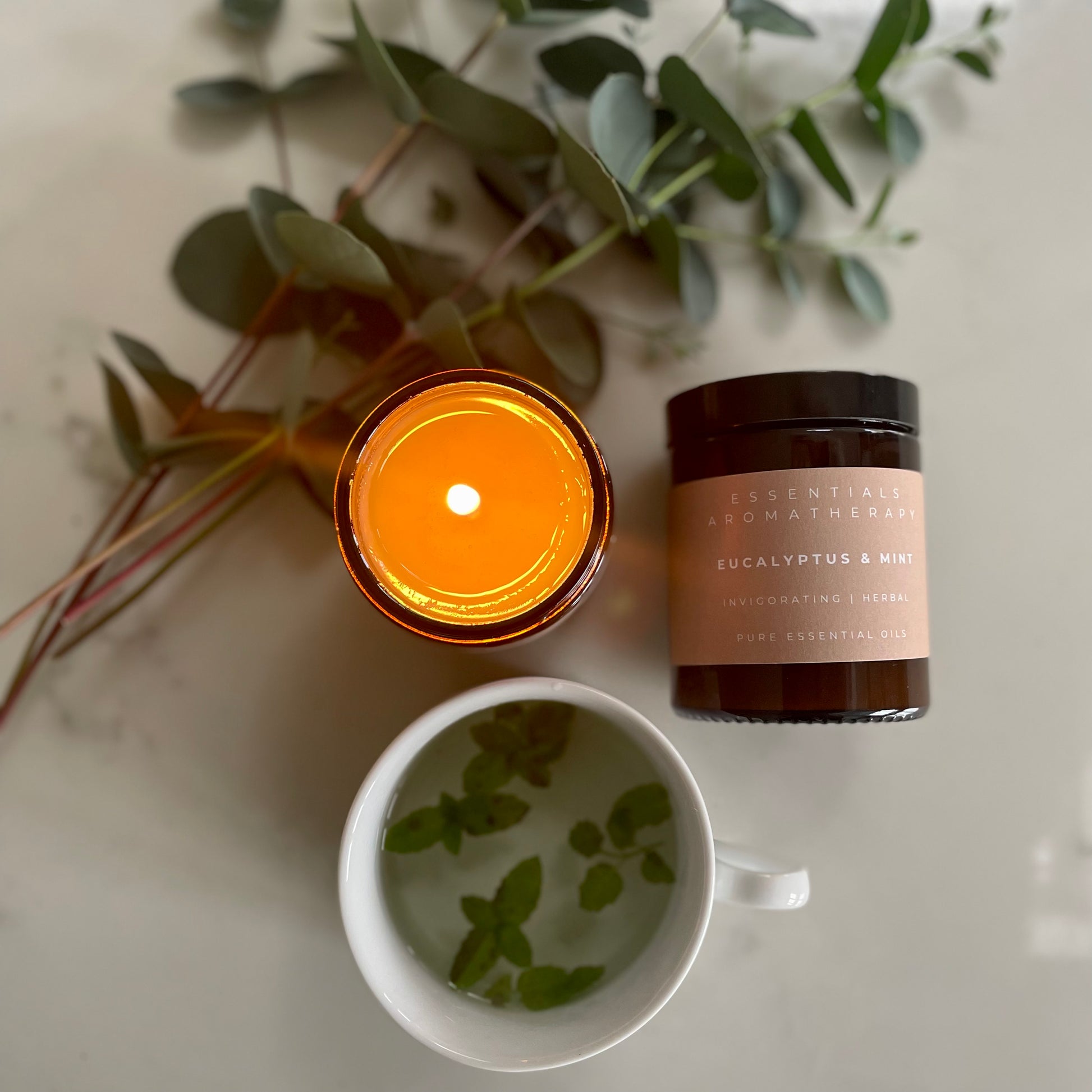 Lit Eucalyptus & Mint essential oil candle beside a cup of mint leaves and fresh eucalyptus branches.